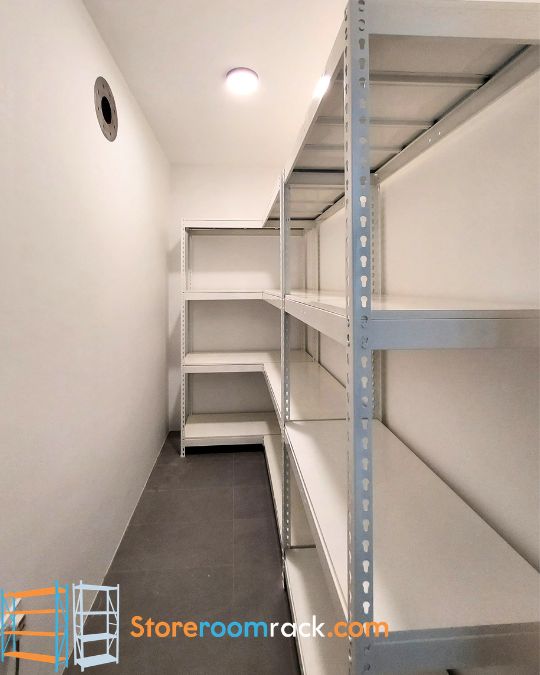 L-shaped boltless rack running the length of an HDB household shelter and turning onto the end wall, with the shelter's round ventilation port on the opposite wall