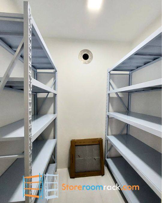 A boltless rack on each of the two long walls of a narrow HDB household shelter, with the round ventilation port on the end wall between them